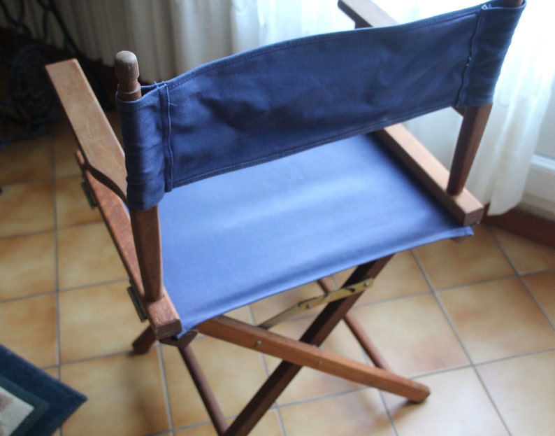 May include: A wooden director's chair with a blue canvas seat and back. The chair is folded and sitting on a tiled floor.