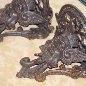 May include: Two ornate, cast iron wall sconces with a weathered, bronze finish. Each sconce features a detailed, classical design with a face, wings, and scrolling elements. The sconces are designed to hold candles or small objects.