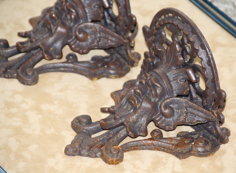 May include: Two ornate, cast iron wall sconces with a weathered, bronze finish. Each sconce features a detailed, classical design with a face, wings, and scrolling elements. The sconces are designed to hold candles or small objects.