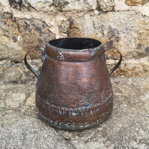 Puede incluir: Una olla de cobre vintage con dos asas. La olla tiene un acabado martillado y una pátina desgastada.