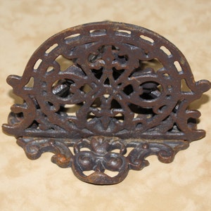 May include: A decorative, cast iron letter holder with an ornate, openwork design. The letter holder is a dark, rusty brown color and features a semi-circular top with a floral and geometric pattern. The base has a stylized, symmetrical design.
