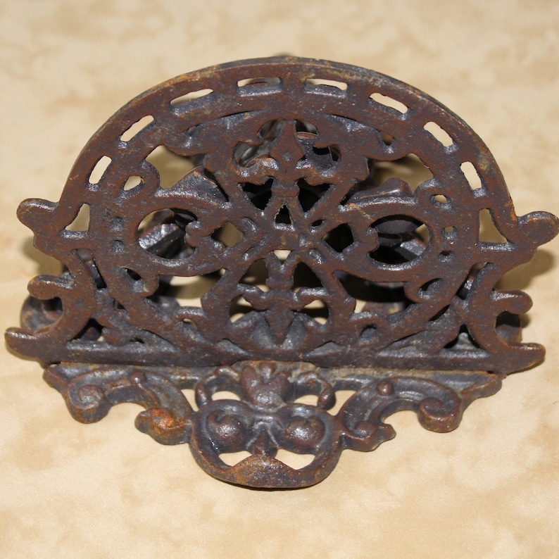 May include: A decorative, cast iron letter holder with an ornate, openwork design. The letter holder is a dark, rusty brown color and features a semi-circular top with a floral and geometric pattern. The base has a stylized, symmetrical design.