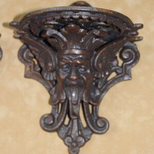 May include: Ornate, dark brown cast iron wall shelf with a decorative face design. The shelf features a circular top and a detailed, stylized face with wings and a beard. The shelf is designed to be mounted on a wall.