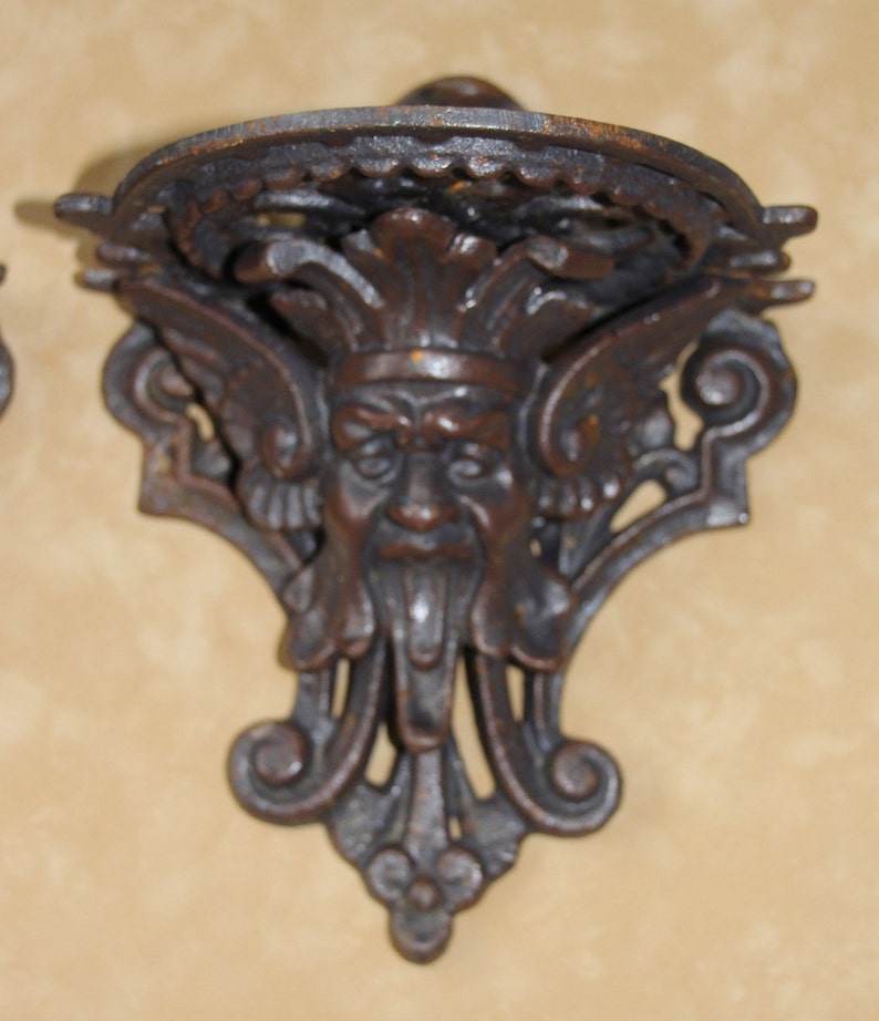 May include: Ornate, dark brown cast iron wall shelf with a decorative face design. The shelf features a circular top and a detailed, stylized face with wings and a beard. The shelf is designed to be mounted on a wall.