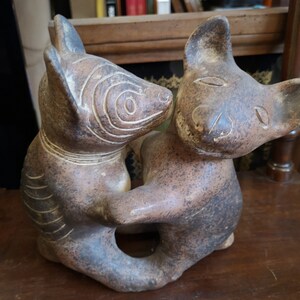 Pode incluir: Duas estatuetas de animais de cerâmica marrom, possivelmente cães, são esculpidas em uma pose brincalhona, com a cabeça de um animal descansando nas costas do outro. As estatuetas têm traços faciais detalhados e superfícies texturizadas.