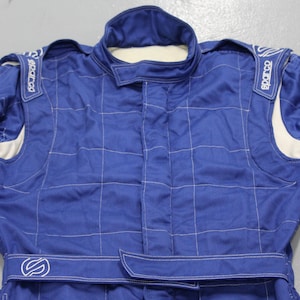 May include: Blue racing suit with white accents and stitching. Features the Sparco logo on the shoulders and a belt with a logo. The suit has a quilted pattern and a high collar.