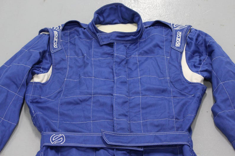 May include: Blue racing suit with white accents and stitching. Features the Sparco logo on the shoulders and a belt with a logo. The suit has a quilted pattern and a high collar.