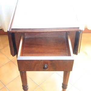 Puede incluir: Una mesa de madera oscura con tapa abatible, un caj&oacute;n y patas torneadas. El caj&oacute;n est&aacute; abierto y la tapa de la mesa es blanca.