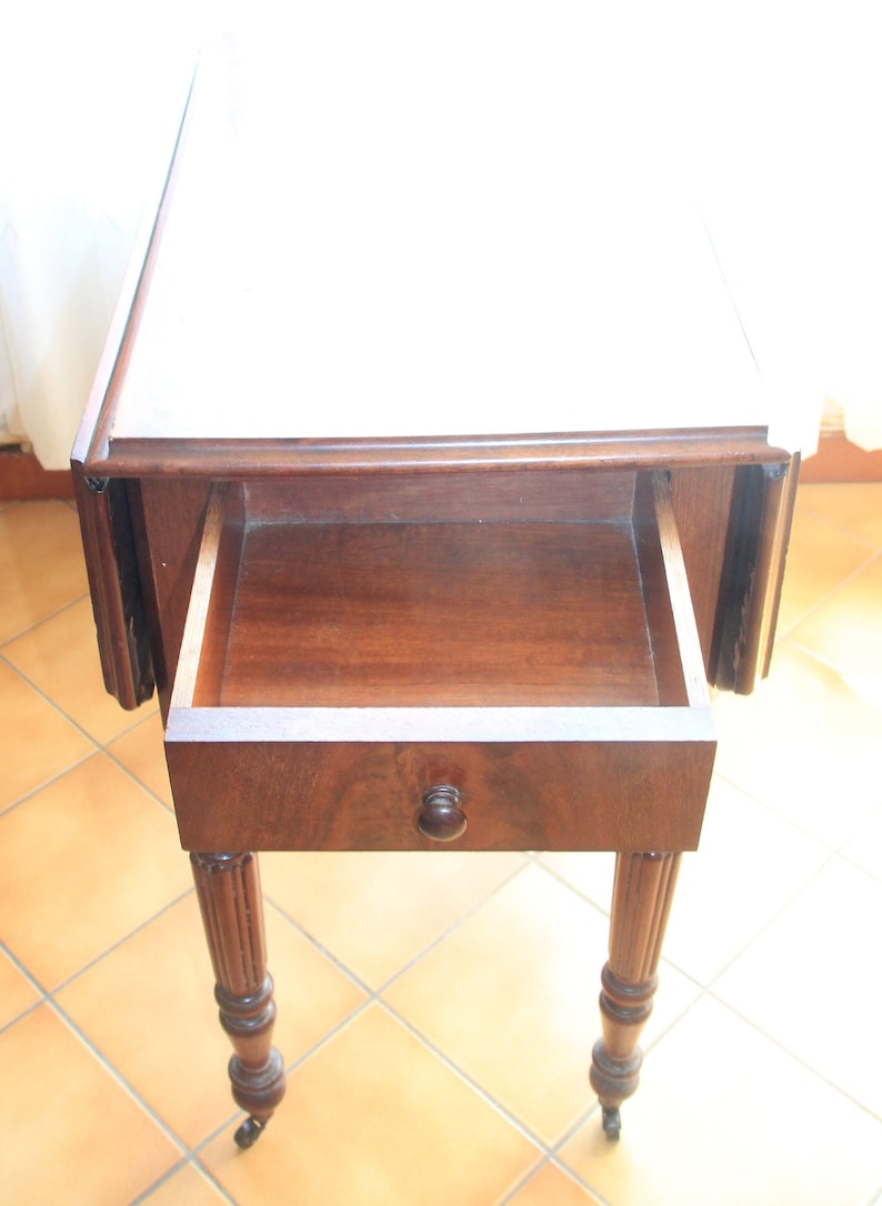 Puede incluir: Una mesa de madera oscura con tapa abatible, un caj&oacute;n y patas torneadas. El caj&oacute;n est&aacute; abierto y la tapa de la mesa es blanca.