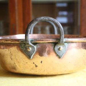 May include: A vintage copper bowl with a hammered finish and a sturdy iron handle. The bowl has a rounded shape and a slightly flared rim. The handle is attached to the bowl with two decorative metal accents.