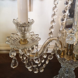May include: A clear glass chandelier with crystal accents. The chandelier features a floral design with hanging crystal droplets and a gold-coloured base. A white candle is in the centre.