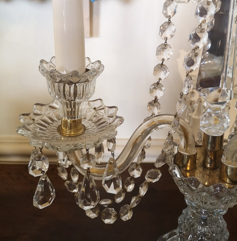 May include: A clear glass chandelier with crystal accents. The chandelier features a floral design with hanging crystal droplets and a gold-coloured base. A white candle is in the centre.