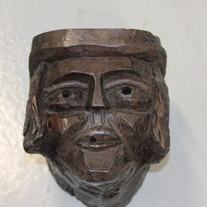 May include: A dark brown, hand-carved wooden sculpture of a human head. The carving features visible chisel marks, a defined brow, almond-shaped eyes with small circular indentations, a straight nose, and a mouth with a small circular opening.