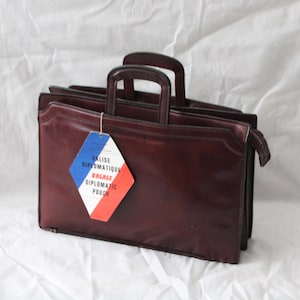 May include: A burgundy leather briefcase with two handles and a zipper closure. A tag with the text "VALISE DIPLOMATIQUE BAGAGE DIPLOMATIC POUCH" in French is attached. The tag has a blue, white, and red design.