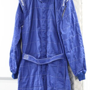 May include: A blue racing suit with white stitching and the Sparco logo on the shoulders. The suit has a high collar, long sleeves, and a belt at the waist. The suit is hanging on a wooden hanger.