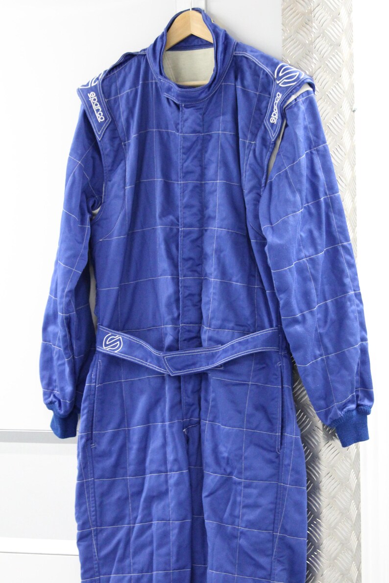 May include: A blue racing suit with white stitching and the Sparco logo on the shoulders. The suit has a high collar, long sleeves, and a belt at the waist. The suit is hanging on a wooden hanger.