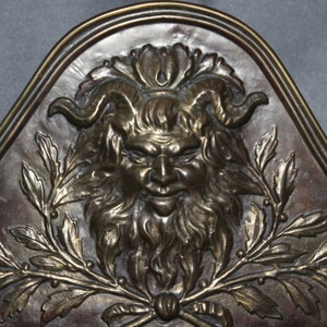 May include: A brass plaque featuring a detailed relief sculpture of a grotesque face with horns and a beard, surrounded by stylised leaves and berries.