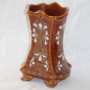 May include: A brown ceramic vase with white and gold floral designs. The vase has a square base with four feet and a flared top.