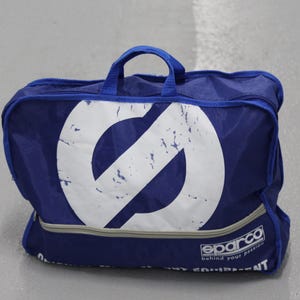 May include: A blue Sparco gear bag with a large white logo on the front. The bag has a handle and a zipper closure. The text "behind your passion" is printed on the bottom.