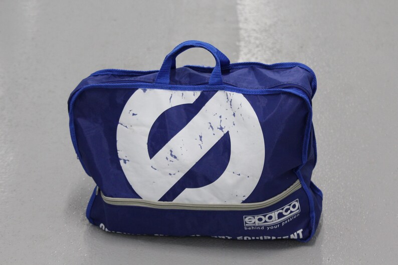 May include: A blue Sparco gear bag with a large white logo on the front. The bag has a handle and a zipper closure. The text "behind your passion" is printed on the bottom.