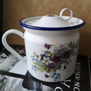 May include: White enamelware mug with a blue rim and a floral design featuring purple and blue flowers. The mug has a lid with a small handle.