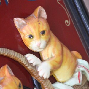 Może przedstawiać: Tablica w stylu vintage z czerwonym tłem i złotymi literami. Na tablicy przedstawiono dwa ceramiczne koty w kolorze pomarańczowym i białym, jeden trzymający linę, a drugi siedzący w koszyku.