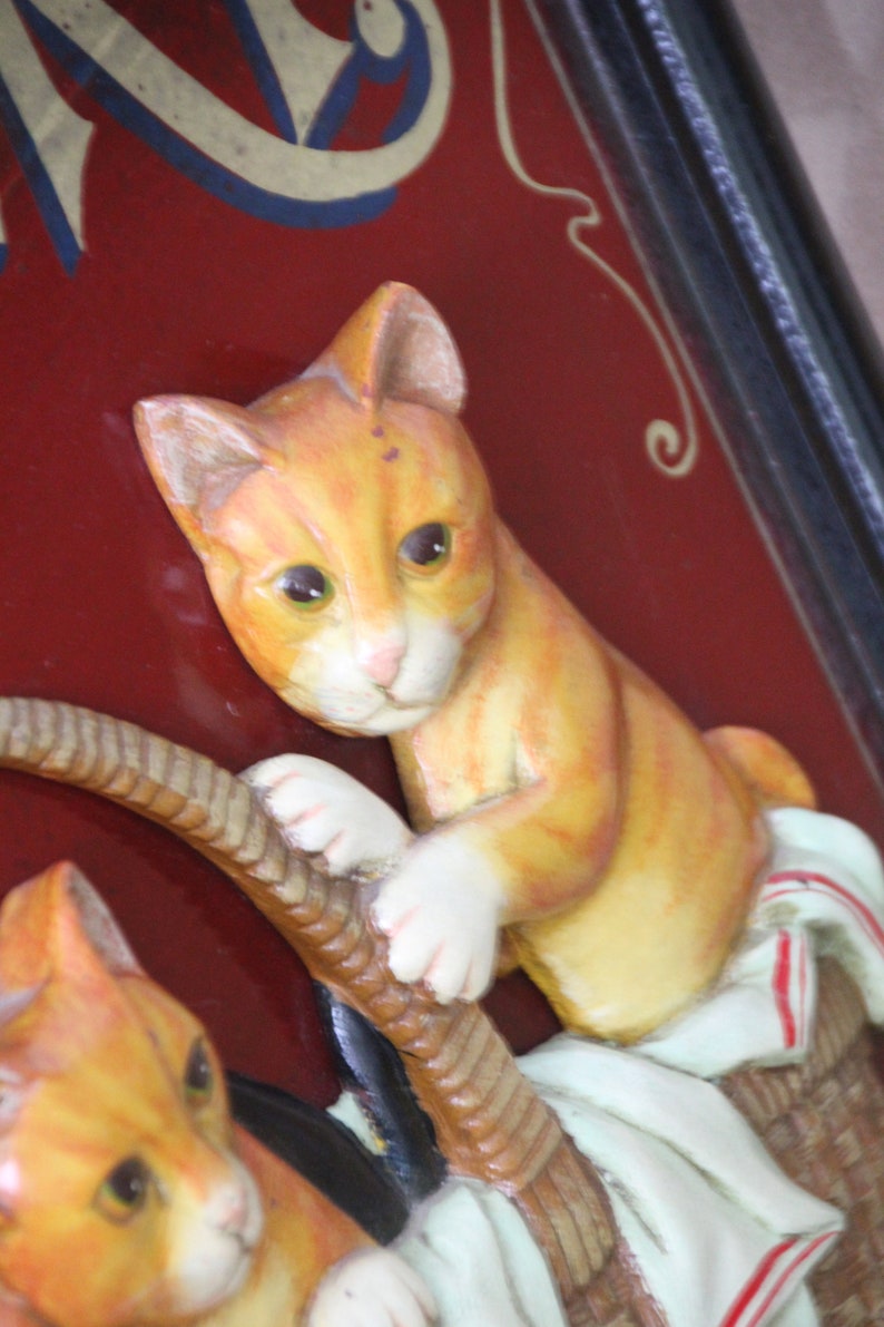 Może przedstawiać: Tablica w stylu vintage z czerwonym tłem i złotymi literami. Na tablicy przedstawiono dwa ceramiczne koty w kolorze pomarańczowym i białym, jeden trzymający linę, a drugi siedzący w koszyku.