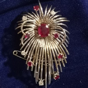 Puede incluir: Un broche de plata con un gran rubí rojo en el centro y rubíes rojos más pequeños y diamantes de imitación transparentes a su alrededor. El broche tiene la forma de una explosión de luz o un fuego artificial.