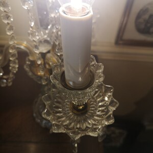 May include: A close-up of a crystal chandelier with a lit bulb. The chandelier has clear, faceted crystal elements and a gold-coloured metal frame. The bulb is encased in a white, candle-shaped sleeve, and the light is bright.