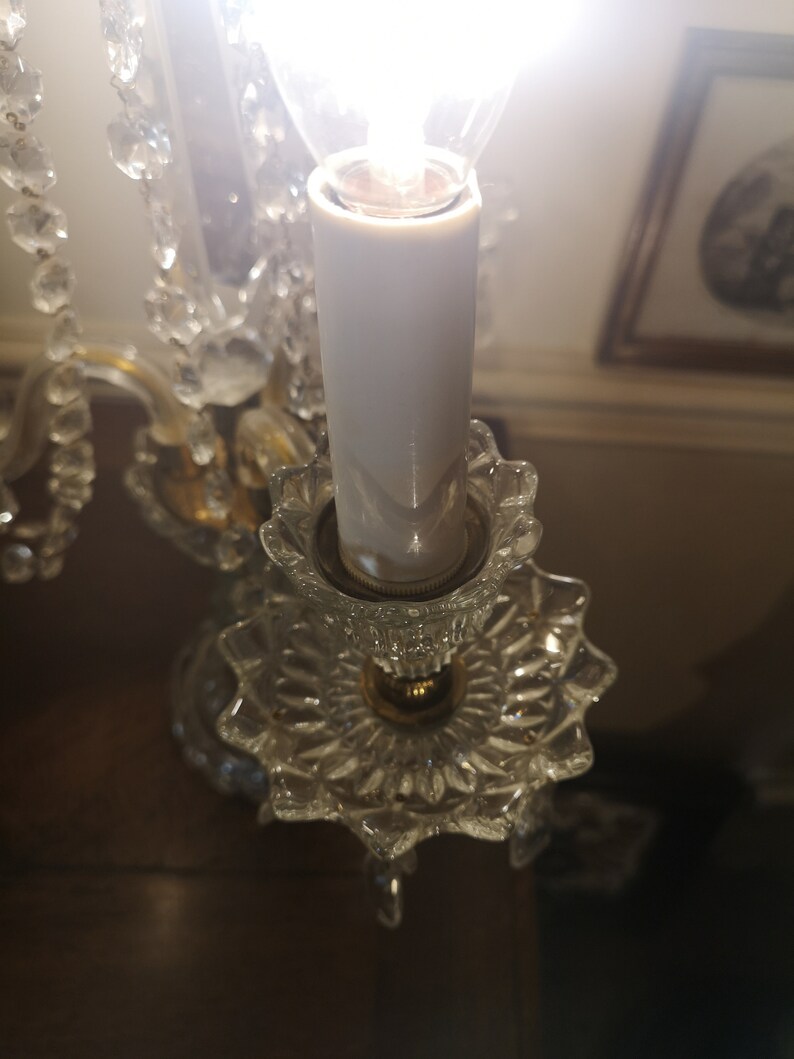 May include: A close-up of a crystal chandelier with a lit bulb. The chandelier has clear, faceted crystal elements and a gold-coloured metal frame. The bulb is encased in a white, candle-shaped sleeve, and the light is bright.