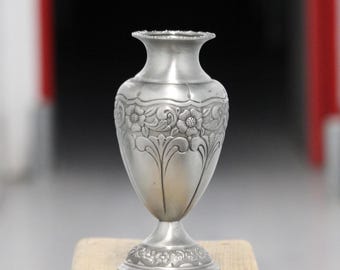May include: A silver vase with a flared rim and a pedestal base. The vase has an embossed floral design around the body. The vase is approximately 18 cm tall.