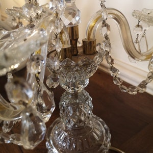 May include: A clear crystal chandelier with multiple arms and hanging crystal accents. The chandelier has a detailed, ornate design with a brass base and a decorative glass stem. The crystals reflect light, creating a sparkling effect.