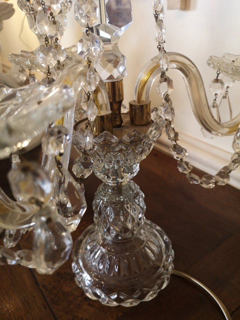 May include: A clear crystal chandelier with multiple arms and hanging crystal accents. The chandelier has a detailed, ornate design with a brass base and a decorative glass stem. The crystals reflect light, creating a sparkling effect.