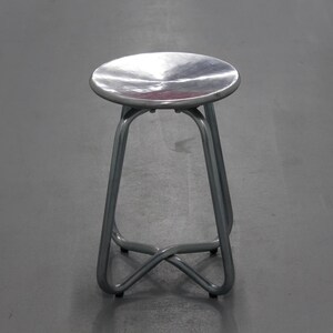 May include: A silver metal stool with a circular seat and curved legs. The legs are joined by a crossbar near the base. The stool is designed for seating.