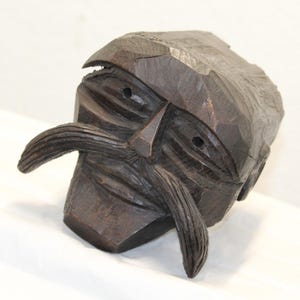 May include: A dark brown hand-carved wooden sculpture of a human head featuring a prominent, long, downward-curving mustache, deep-set eyes, and a flat, angular forehead. The surface displays visible chisel marks and a rough, textured finish.