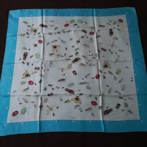 May include: A white silk scarf with a blue border and a pattern of various insects, including beetles, butterflies, and ants. The scarf has the text "HERMES PARIS" in the center.