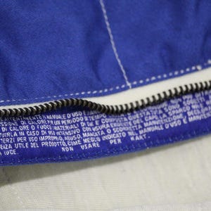 May include: Close-up of a blue fabric with a white zipper and white stitching. The fabric has a label with white text. The text includes the words "offer", "wear", and "kart".