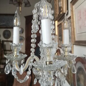 May include: A vintage crystal chandelier with three candle-shaped lights. The chandelier features clear crystal accents, including hanging teardrop-shaped crystals. The arms are a light gold colour. The background includes framed artwork.