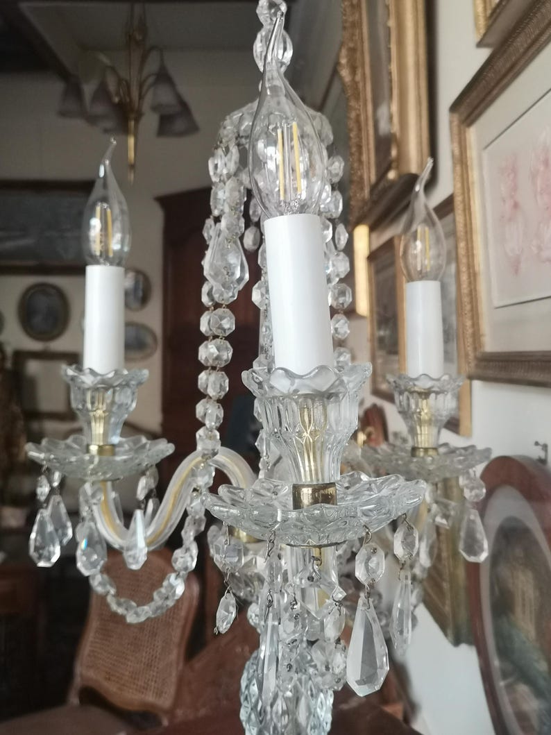 May include: A vintage crystal chandelier with three candle-shaped lights. The chandelier features clear crystal accents, including hanging teardrop-shaped crystals. The arms are a light gold colour. The background includes framed artwork.