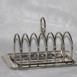 May include: A silver-toned metal toast rack featuring six arched slots for holding slices of bread. The rack has a rectangular base with beveled edges and a central circular handle loop at the top for carrying. The finish is smooth and reflective.