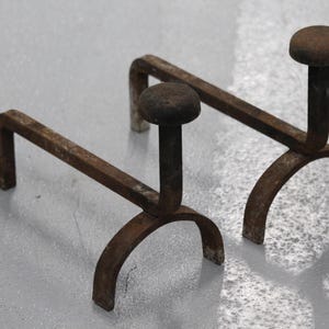 Puede incluir: Un par de morillos de chimenea a juego fabricados en metal de color marrón oscuro desgastado. Cada morillo presenta una base arqueada, una barra de soporte horizontal y un poste vertical rematado con un disco circular plano para leña.