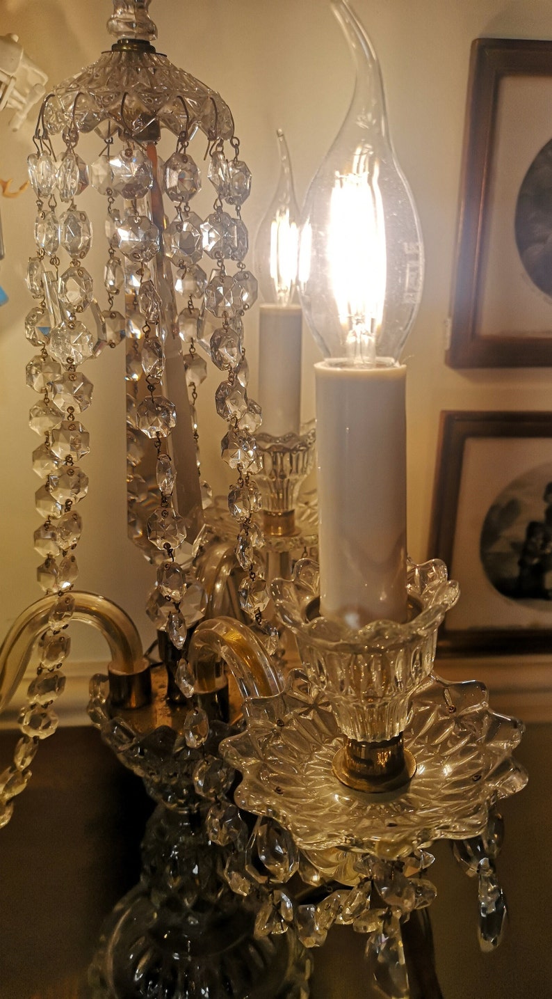 May include: A vintage crystal chandelier with multiple candle-shaped light bulbs. The chandelier features hanging crystal accents and ornate glass detailing. The light bulbs are lit, casting a warm glow. The fixture is likely made of glass and metal.
