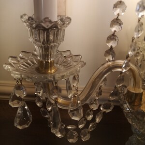May include: Close-up of a crystal chandelier with a white candle. The chandelier features clear, faceted crystal pendants and strands, with a gold-coloured metal frame. The candle holder has a decorative, scalloped edge.