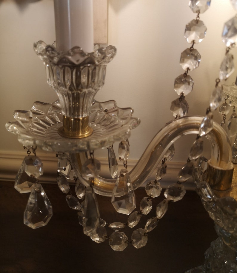 May include: Close-up of a crystal chandelier with a white candle. The chandelier features clear, faceted crystal pendants and strands, with a gold-coloured metal frame. The candle holder has a decorative, scalloped edge.