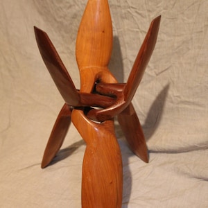 May include: A handcrafted wooden sculpture with a light brown finish. The sculpture features a central vertical element with three outward-extending, curved arms. The arms are connected to a base, creating a unique, abstract design.
