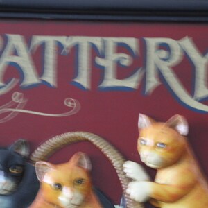 Może przedstawiać: Czerwony znak z napisem "CATTERY" w złotych literach. Znak ma ozdobny wz&oacute;r w postaci zwijanego papieru i przedstawia trzy figurki kot&oacute;w, jednego czarnego i dwa pomarańczowe.