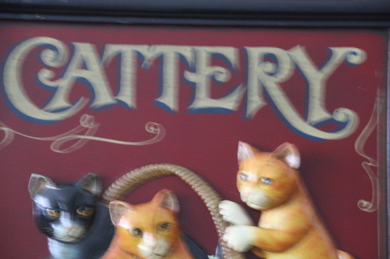 Może przedstawiać: Czerwony znak z napisem "CATTERY" w złotych literach. Znak ma ozdobny wz&oacute;r w postaci zwijanego papieru i przedstawia trzy figurki kot&oacute;w, jednego czarnego i dwa pomarańczowe.