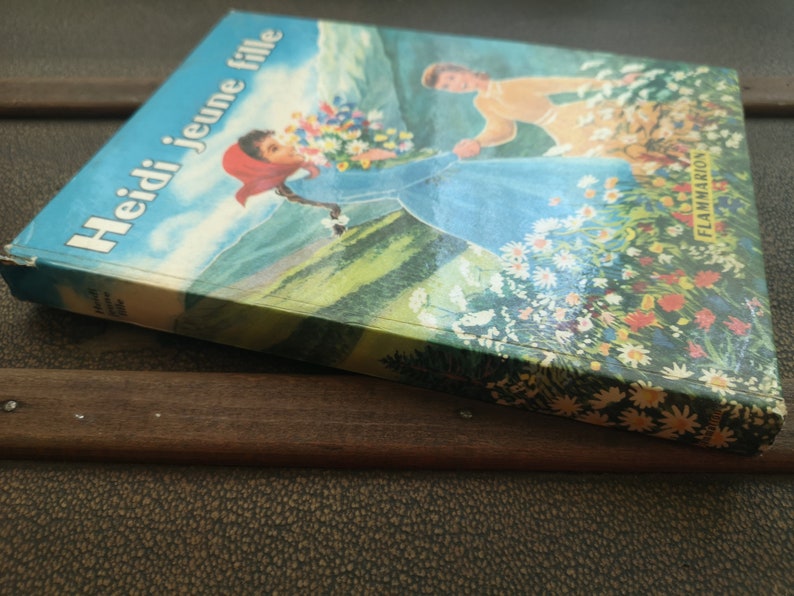 May include: A vintage book titled "Heidi jeune fille" by Flammarion, featuring a colorful illustration of a girl in a blue dress walking through a field of flowers.