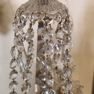 May include: Close-up of a crystal chandelier with multiple strands of faceted crystal beads. The chandelier has a decorative top and a brass-coloured metal base. The crystals reflect light, creating a sparkling effect.