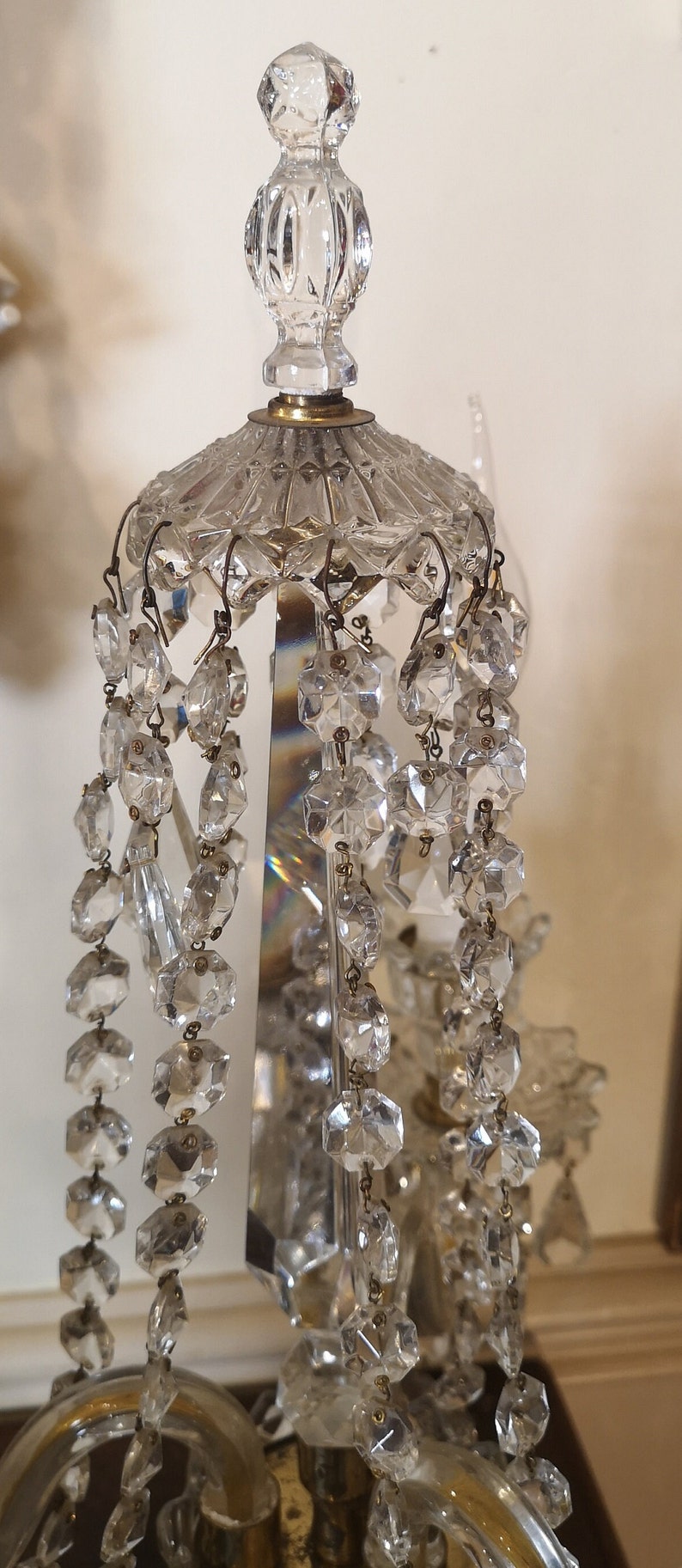 May include: Close-up of a crystal chandelier with multiple strands of faceted crystal beads. The chandelier has a decorative top and a brass-coloured metal base. The crystals reflect light, creating a sparkling effect.